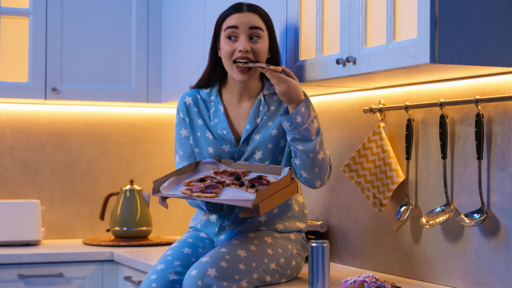 Image of a woman in pajamas snacking at night on pizza